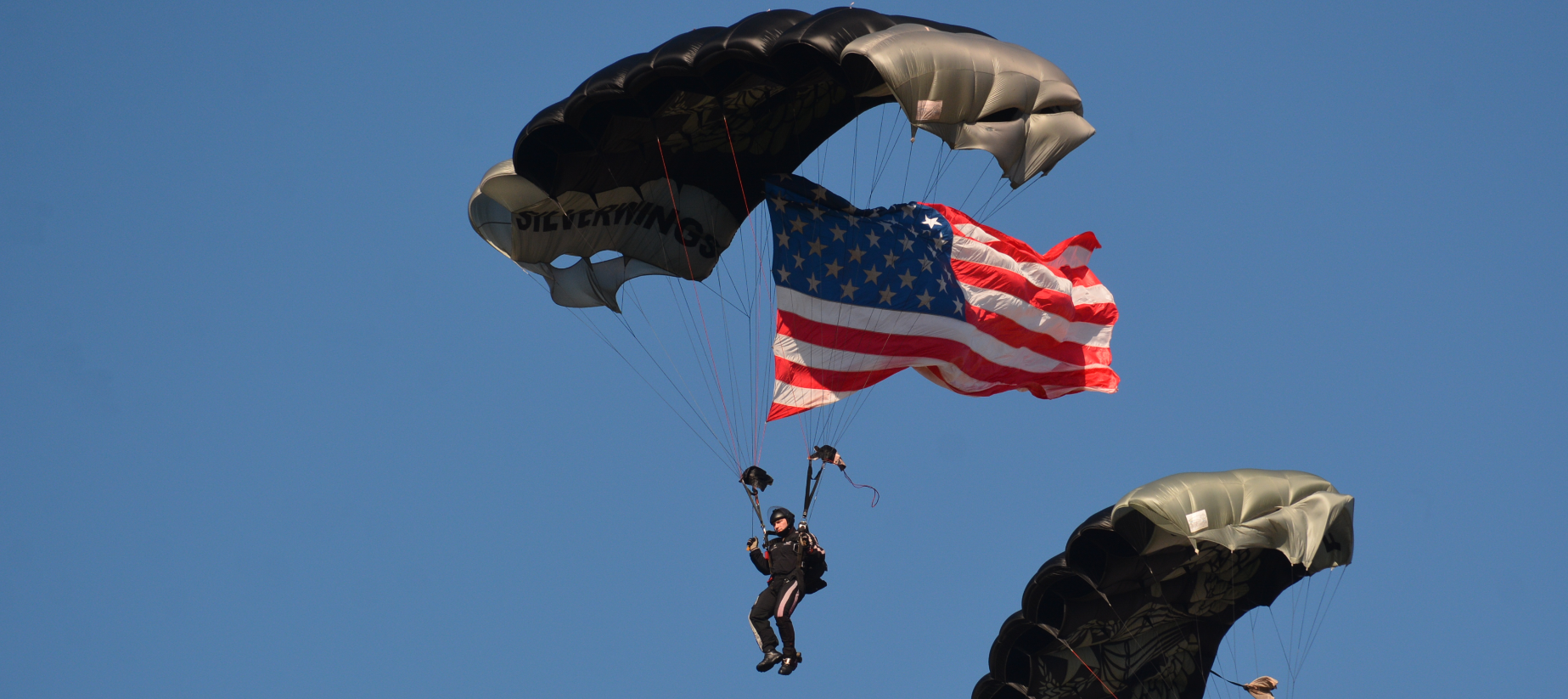 A parachutist descends through the sky carrying an American flag, with another parachutist partially visible to the right.