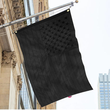 A Jetlifee 3×5 FT Embroidery All Black American Flag Made in USA, crafted from durable polyester and featuring embroidered stars, is mounted on a pole attached to the facade of a building, with another building visible in the background.