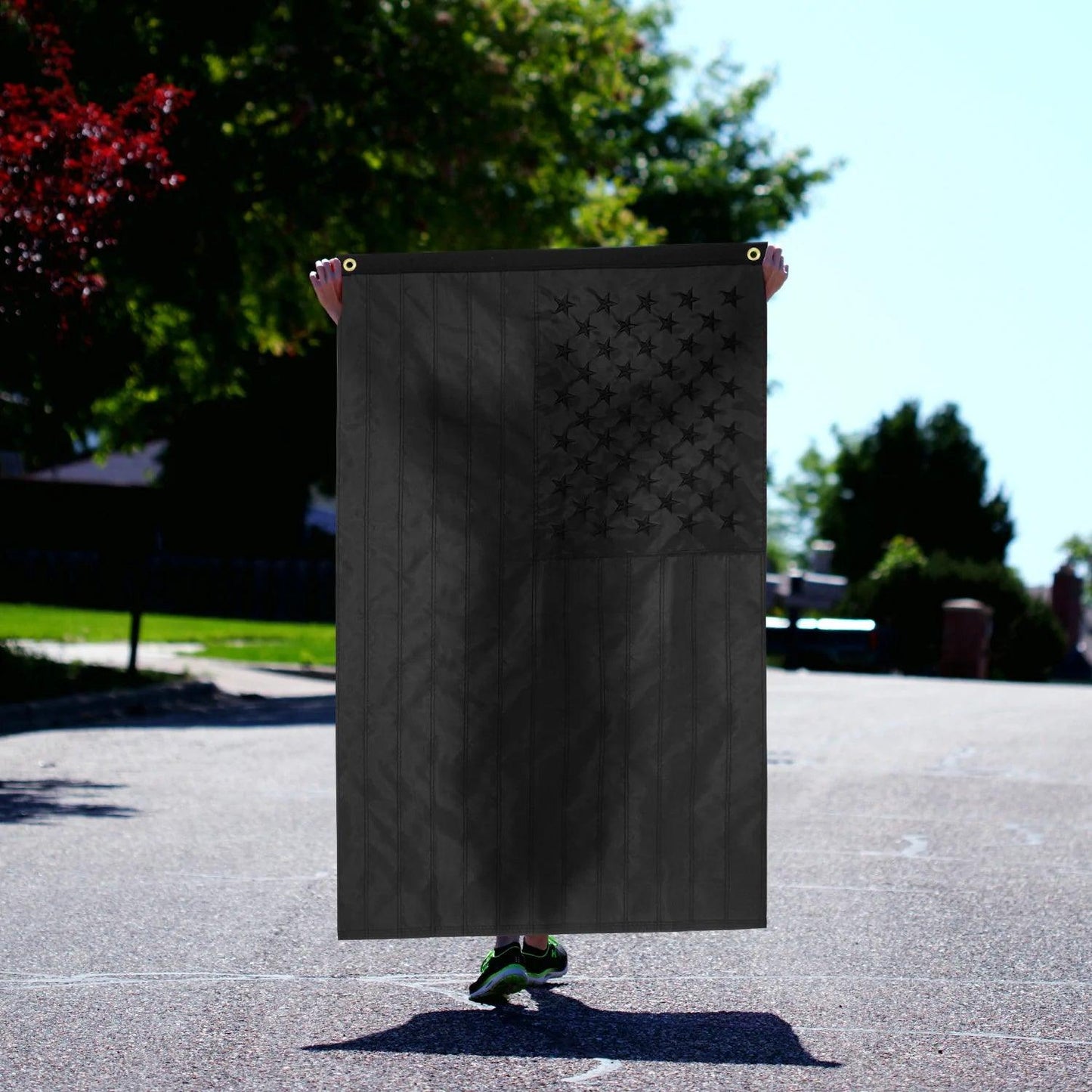 A person is holding a 3×5 FT Embroidery All Black American Flag Made in the USA by Jetlifee, standing in the middle of a suburban street. Only their hands and feet are visible. Trees and houses serve as a backdrop to this durable polyester flag fluttering proudly.