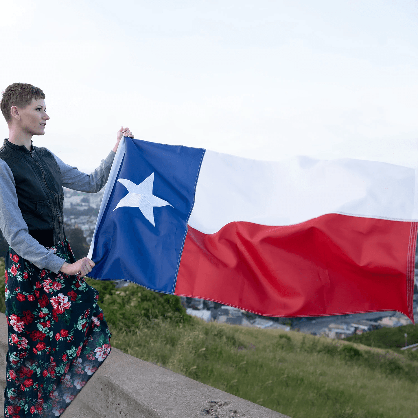 A person stands outdoors holding a 3×5 Ft Embroidery Texas State Flag Made in USA by Jetlifee, crafted from durable heavyweight polyester with brass grommets, the cityscape serving as a striking backdrop.