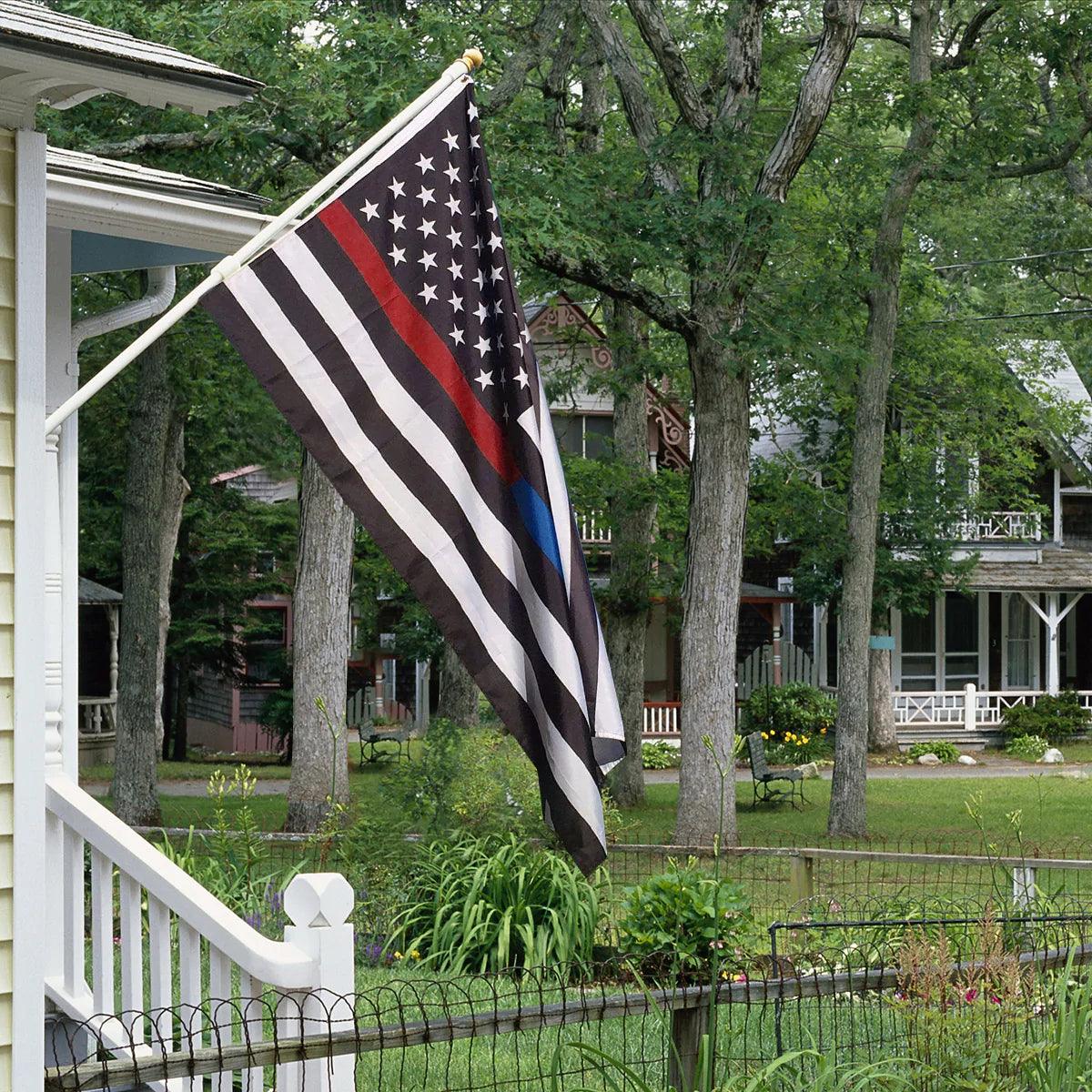 A Jetlifee "3×5 Ft Embroidery Thin Blue Line Thin Red Line Flag Made in USA," featuring black, white, and gray stripes with a red and blue stripe symbolizing police enforcement, is displayed on a house porch in a residential neighborhood. Made from durable and strong heavyweight nylon, this flag stands resilient against the elements.