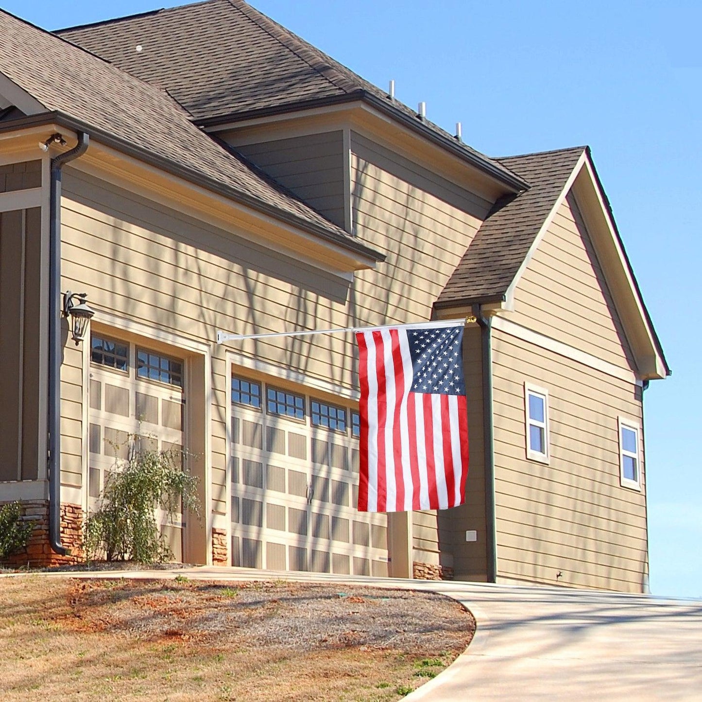 A beige house with three garage doors displays an American flag on a Jetlifee 6ft Aluminum Tangle Free Spinning Flagpole and Multi Position Mounting Bracket Set extending from the front wall. The driveway is concrete, and the sky is clear.