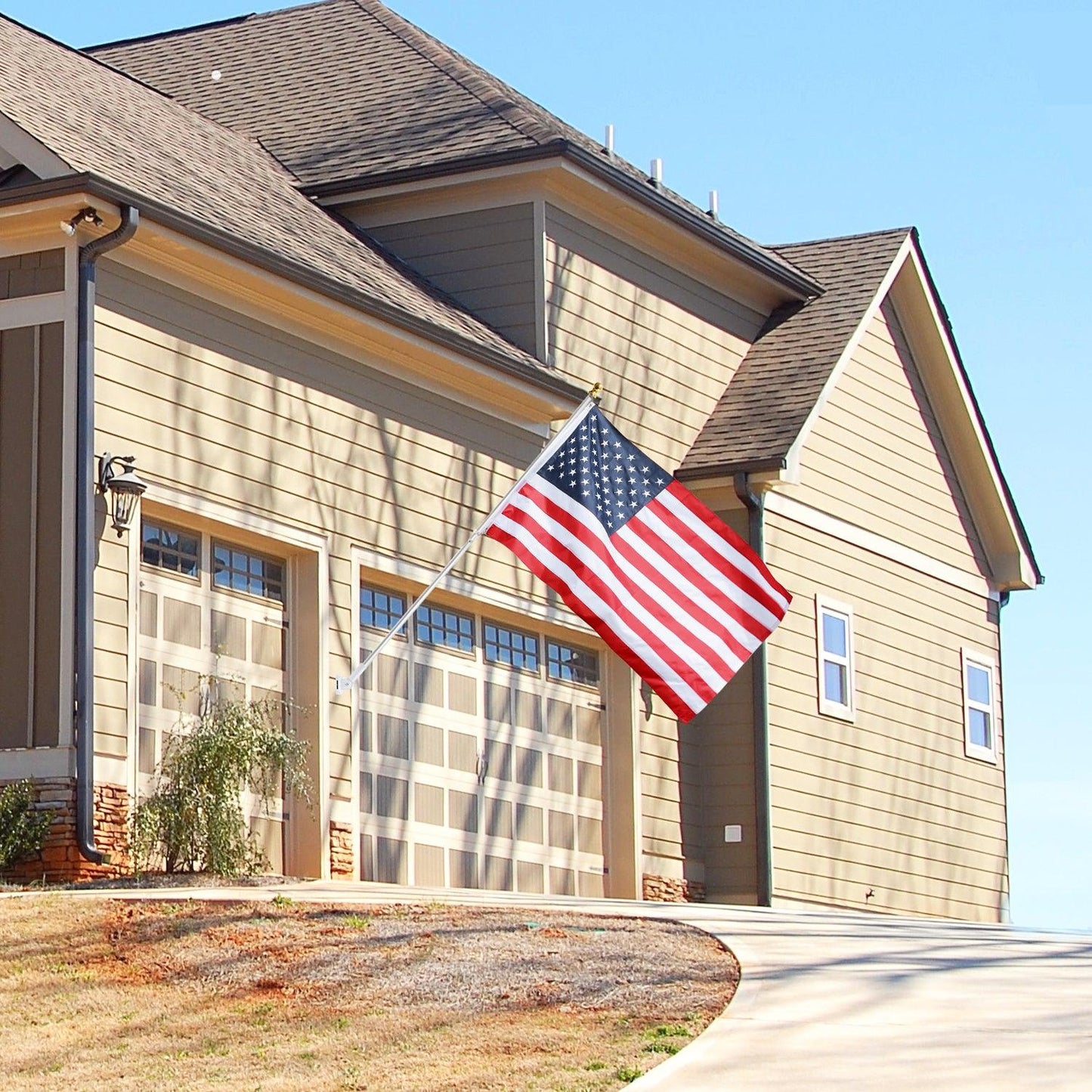 A beige house with an attached garage, displaying an American flag mounted on a Jetlifee 6ft Aluminum Tangle Free Spinning Flagpole and Multi Position Mounting Bracket Set near the entrance, and a clear sky in the background.