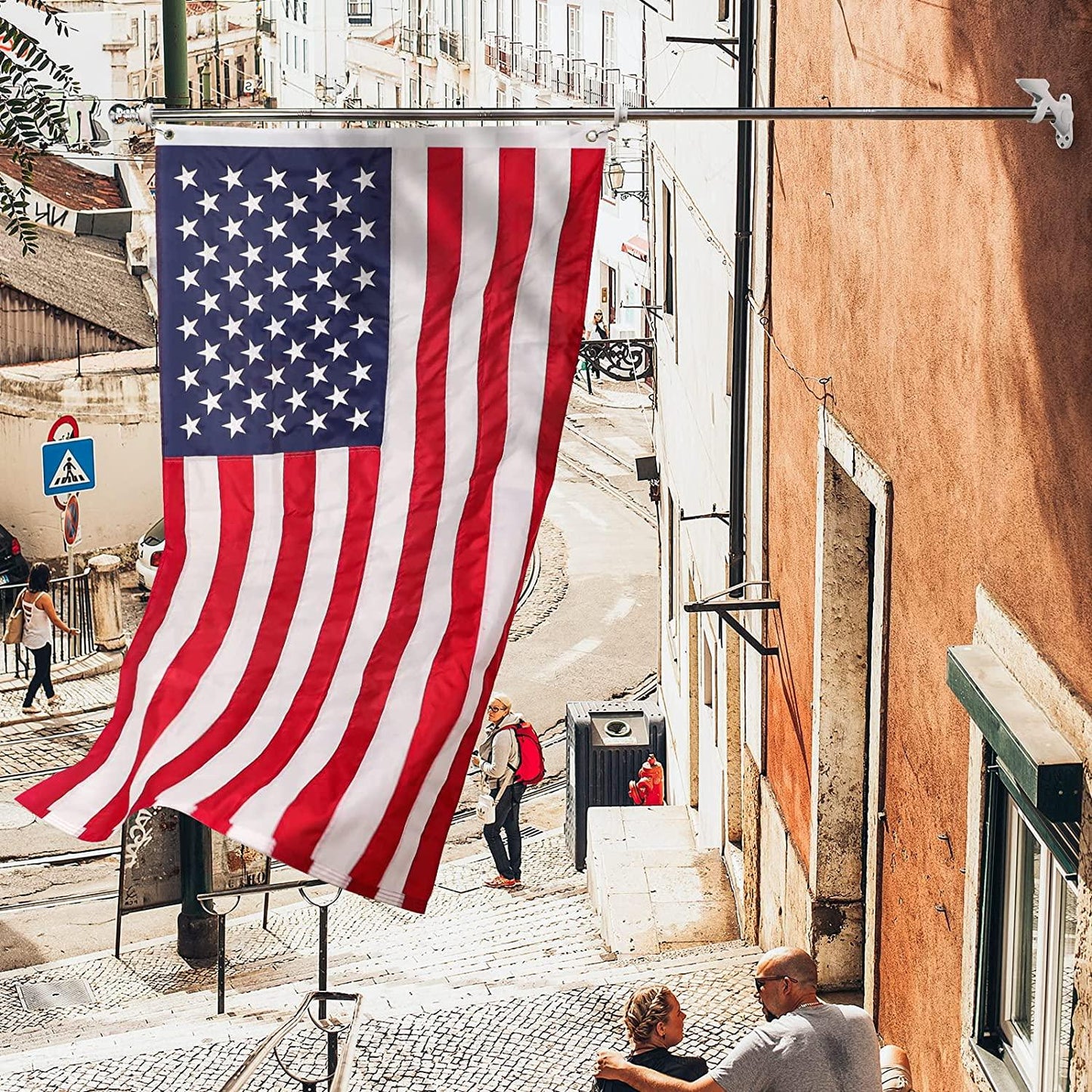 A large American flag, from the Jetlifee American Flag Pole Kit Made in USA with a 6FT 5 Section Flag Pole, hangs from a building on a narrow, cobblestone street with pedestrians walking and talking in the background.