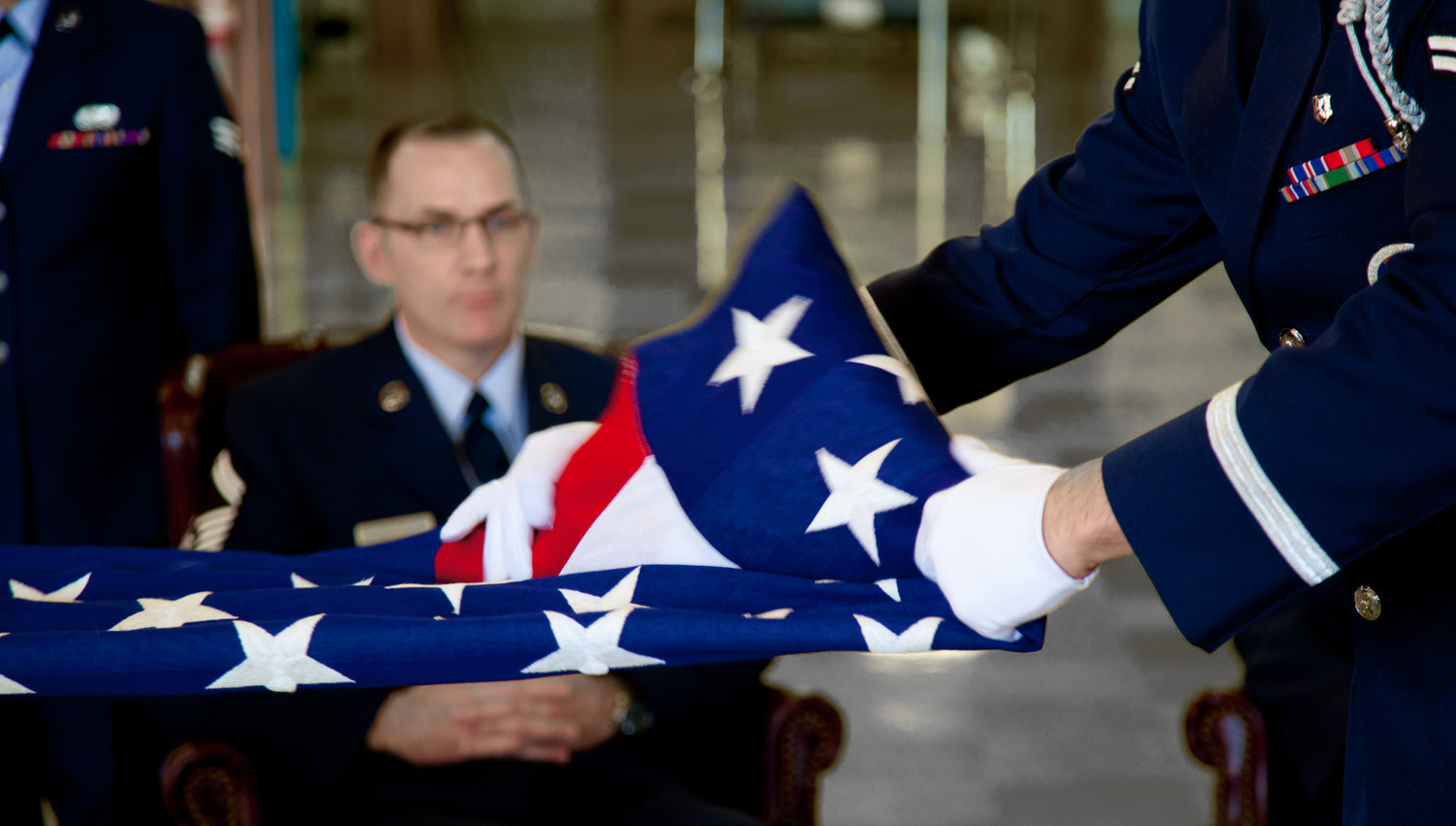 Service members in uniform ceremoniously fold an American flag; one seated in the background and another handling the flag in the foreground.