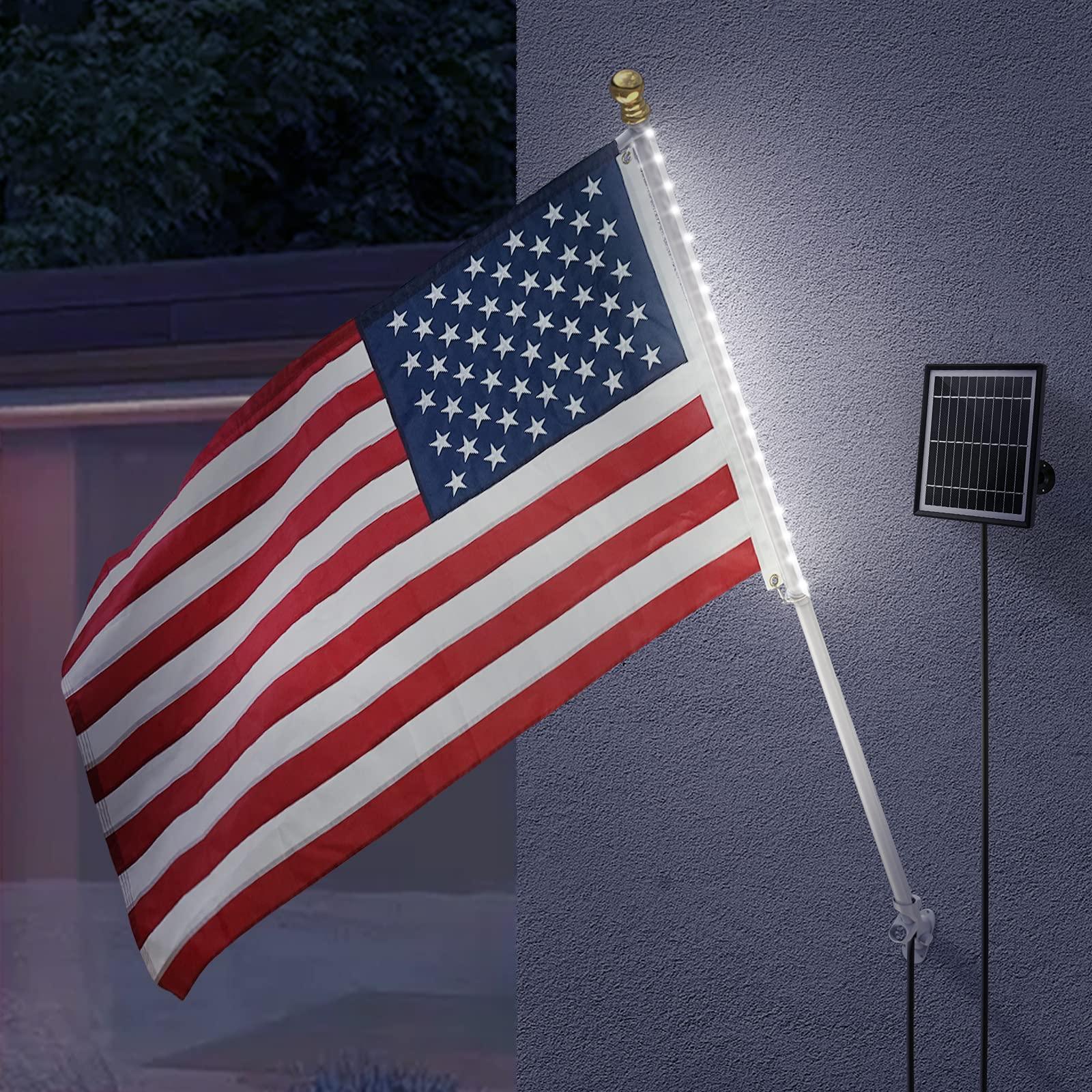 A 3x5 USA flag from the Jetlifee Solar American Flag Pole Light Kit, manufactured by Jetlifee, is mounted on a wall with an attached LED light strip. A nearby small solar panel powers the lights, creating a solar-powered display.